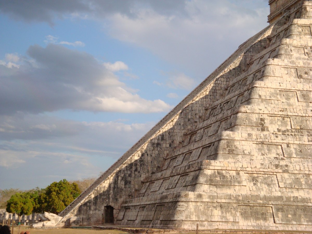 Limestone pyramid with a triangular shadow pattern cast on one side of the pyramid where the remaining sunlight resembles the body of a snake connected to the carved head of a serpent at the base of the pyramid