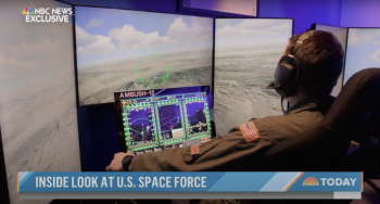 Man sitting at desk looking at a laptop and two large screens with a photo of land from high up in the air