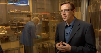 Man in a suit stands in front of a clean room with two workers.