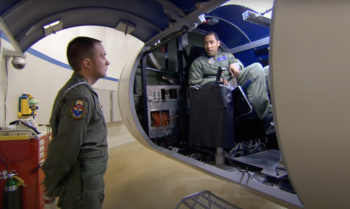 Two service members one standing on the ground one sitting in a flight simulator.