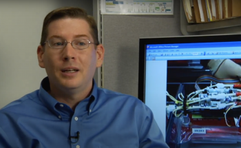 Man on left of screen with a computer screen behind him.