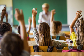 A classroom of children with their hands raised looking at the teacher