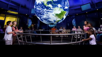 Photo of a room of students with a model Earth suspended above them and text that says Science on a Sphere