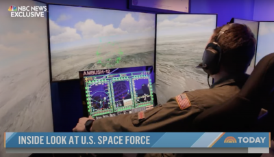 Man sitting at desk looking at a laptop and two large screens with a photo of land from high up in the air
