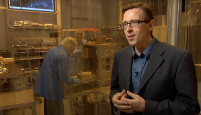 Man in a suit stands in front of a clean room with two workers.