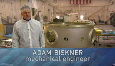 Man in a white coat in front of a large round piece of meatal with text saying Adam Biskner mechanical engineer