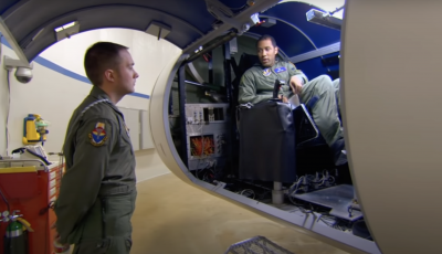 Two service members one standing on the ground one sitting in a flight simulator.