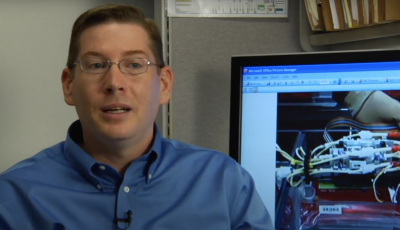 Man on left of screen with a computer screen behind him.