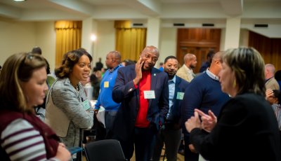 Participants stand around a table, listening to one speaker intently.