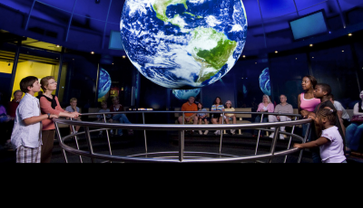 Photo of a room of students with a model Earth suspended above them and text that says Science on a Sphere