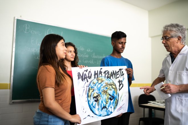 Three students stand holding a sign with a painted globe on it that says "there is no planet B" in Portuguese.