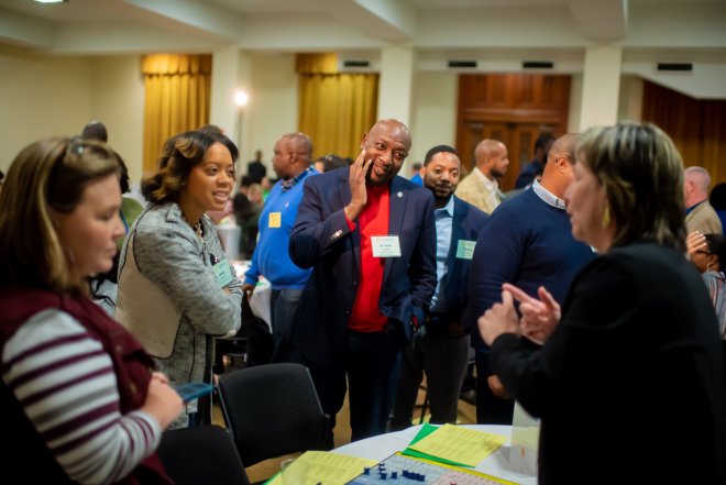 Participants stand around a table, listening to one speaker intently.